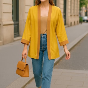 Op de afbeelding: Een vrouw draagt een mosterdgele jas met bruine biezen, een beige top en een blauwe spijkerbroek. Ze draagt een bruine handtas en bruine hakken. De outfit is geschikt voor een informele of semi-formele gelegenheid.