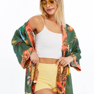Pode incluir: Um quimono verde com um design floral e tropical vibrante, com detalhes em laranja e amarelo. O quimono é combinado com um top branco e calções amarelos. A pessoa está usando óculos de sol amarelos.
