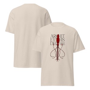 Peut inclure: T-shirt beige avec un motif rouge et noir de calmar géant et le texte "ARCHITEU DUX GIANT SQUID". Le t-shirt a un col rond et des manches courtes. Le dos du t-shirt est beige uni.