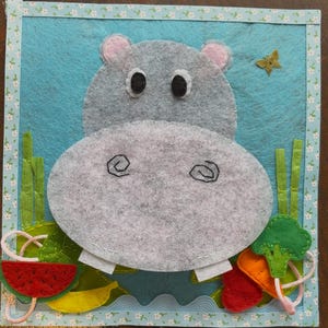 May include: A handmade felt hippo toy with a light blue background and a decorative border. The hippo is gray with pink ears and black eyes. It has a felt mouth with white teeth and is surrounded by felt fruits and vegetables.