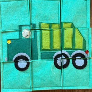Pu&ograve; includere: Un puzzle in feltro verde raffigurante un camion. Il camion ha un conducente, ruote nere e un bottone giallo. Il puzzle &egrave; composto da diversi pezzi quadrati su uno sfondo di legno.