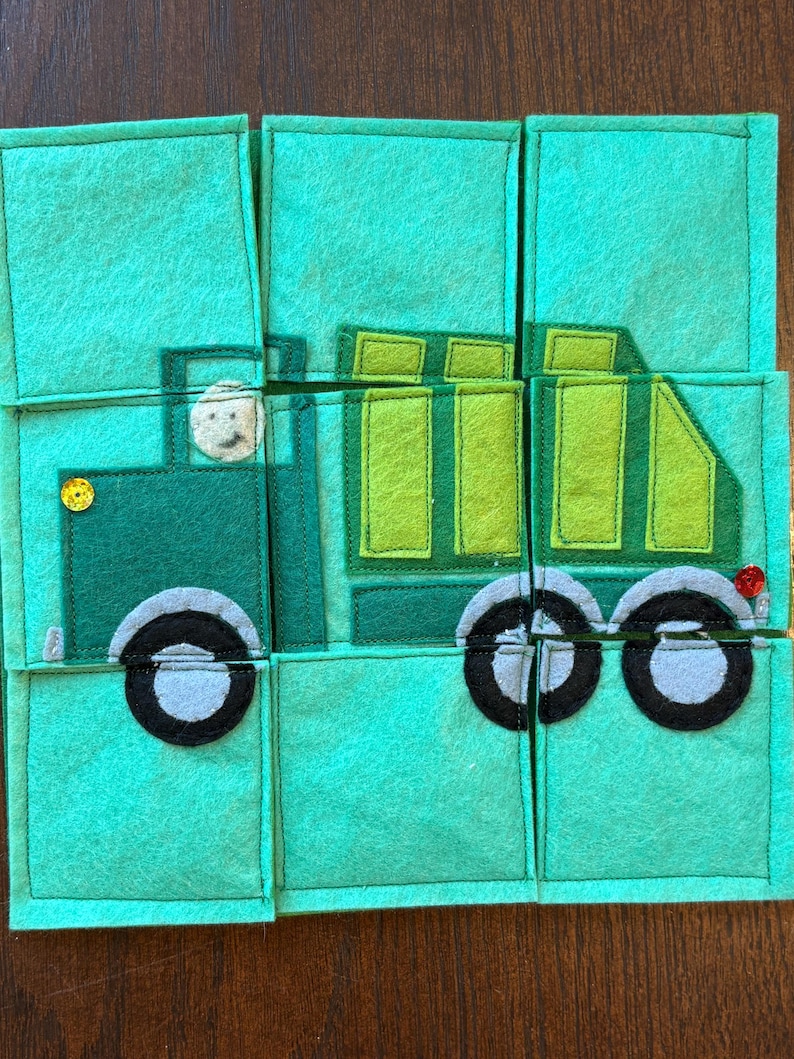 Pu&ograve; includere: Un puzzle in feltro verde raffigurante un camion. Il camion ha un conducente, ruote nere e un bottone giallo. Il puzzle &egrave; composto da diversi pezzi quadrati su uno sfondo di legno.