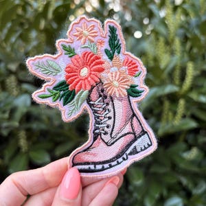 May include: Embroidered patch featuring a pink boot filled with a bouquet of flowers. The boot is light pink with black laces and a white sole. The flowers are in shades of coral, peach, and pink, with green leaves. The patch has a pink border.