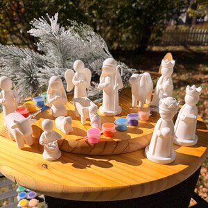 May include: A set of white ceramic nativity figurines including figures of people, animals, and a manger. The set is accompanied by small circular containers of colorful paint and a thin paintbrush, designed for painting the unpainted white surfaces.