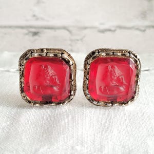 May include: A pair of vintage cufflinks featuring square, ruby-red glass stones with a carved profile design. The stones are set in ornate, silver-toned metal frames. The cufflinks are displayed on a white, textured surface.