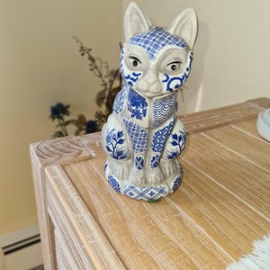May include: A ceramic cat figurine featuring a white base with intricate blue patterns, including floral motifs, cross-hatching, and geometric designs covering the ears, face, chest, and legs. The cat is in a seated position with its tail curled.
