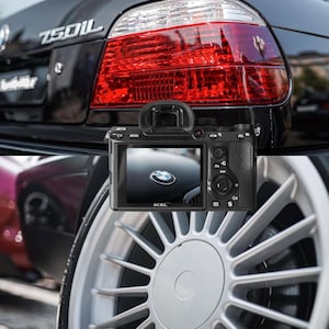 Può includere: Primo piano della parte posteriore di un'auto nera con fanale rosso e fotocamera digitale. Lo schermo della fotocamera mostra il cofano di un'auto con il logo BMW. Un cerchio in lega d'argento è in primo piano.