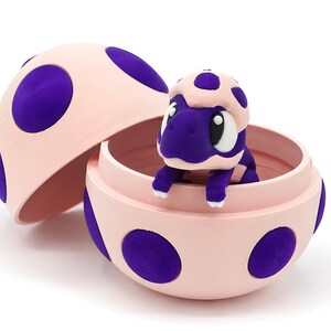 Puede incluir: Un juguete de tortuga morada sale de un recipiente rosa en forma de huevo con lunares morados. La tapa del huevo está abierta, revelando la tortuga en su interior. El juguete tiene detalles blancos.