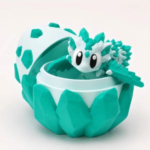 Puede incluir: Una figura de criatura de fantasía turquesa y blanca que emerge de un contenedor a juego en forma de huevo. El contenedor tiene un diseño geométrico, y la criatura tiene ojos grandes y detalles puntiagudos.