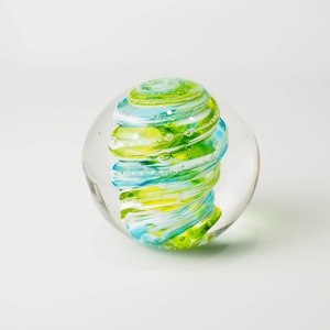 May include: A clear glass paperweight with a swirling pattern of green, blue, and white glass inside.