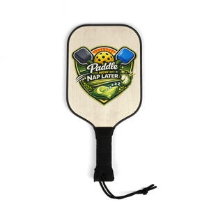 Peut inclure: Une raquette de pickleball avec une face en bois et une poignée noire. La raquette présente un graphisme coloré avec le texte "Paddle Now Nap Later", une balle de pickleball et deux raquettes.