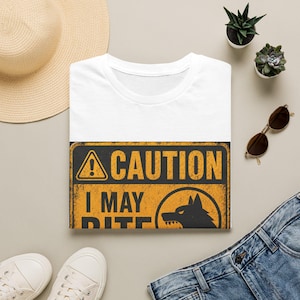 以下が含まれることがあります： 「CAUTION I MAY BITE」の文字とオオカミのシルエットが描かれた、黒と黄色のグラフィックが特徴の白いTシャツ。Tシャツは、麦わら帽子、サングラス、ジーンズと一緒に、明るい表面に折りたたまれています。