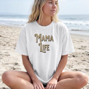 May include: White t-shirt with the text "Mama Life" in a vintage-style font. The text is in a combination of gold and black. The shirt is a casual, everyday wear item.