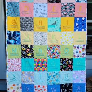 May include: A colorful alphabet quilt with each letter embroidered on a different fabric square. The squares feature various patterns, including animals, food, and geometric designs. The letters are A through Z, with each letter representing a word. For example, A is for Alligator, B is for Bicycle, C is for Clouds, and so on.