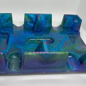 May include: A rectangular resin coaster holder featuring a swirling marbled pattern of deep blue, vibrant turquoise, and hints of purple. The structure includes six upright dividers and a central recessed slot, creating a glossy, multi-colored finish.