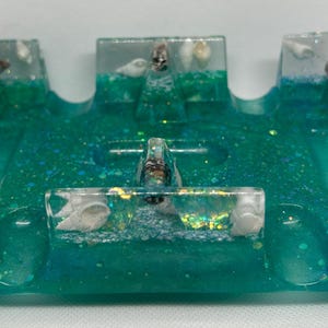 May include: A rectangular teal resin tray featuring embedded white and brown seashells along the raised edges. The translucent material contains fine glitter particles throughout, creating a shimmering effect within the deep blue-green base.