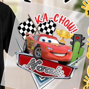 Puede incluir: Una calcomanía de transferencia térmica con un coche de carreras rojo con el número 95, dos banderas a cuadros, un trofeo dorado, un cactus verde y un semáforo. El texto dice KA-CHOW! y el nombre Noah en fuente metálica tipo diamante.