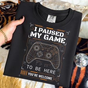 May include: Black sweatshirt with the text "I PAUSED MY GAME TO BE HERE YOU'RE WELCOME" and a white and orange graphic of a video game controller. The sweatshirt is held by a person.