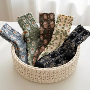 May include: A woven basket filled with six pairs of patterned socks. The socks feature floral and geometric designs in various colors, including blue, green, brown, and cream. The basket is made of a light beige material.