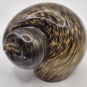 May include: A decorative glass sculpture in the shape of a snail shell. The shell features a swirling pattern of brown and tan hues. The sculpture has a glossy finish and is set against a white background.