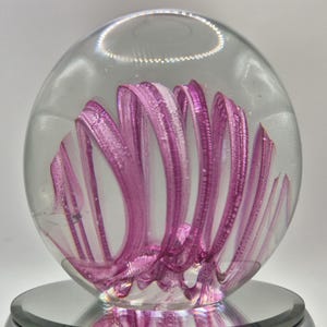 May include: A clear glass paperweight with a swirling, abstract design in shades of pink and white. The design resembles a stylized flower or spiral, encased within the spherical glass. The paperweight sits on a reflective, circular base.