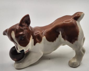 Puede incluir: Figura de cerámica de un perro juguetón, con pelaje marrón y blanco. El perro está representado con la cabeza gacha, sosteniendo una bola marrón oscura en la boca. La figura tiene un acabado brillante.