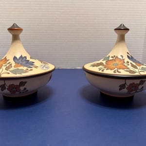 May include: Two matching ceramic lidded jars. The jars are cream-colored with floral designs in orange, blue, and brown. Each jar has a pointed finial and a dark brown rim. The jars are on a blue surface.