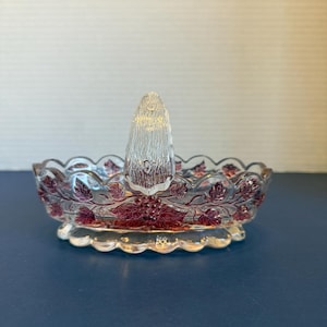 May include: Clear glass ring holder with a raised, textured center and a scalloped edge. The bowl is decorated with a burgundy floral and leaf pattern. The ring holder is designed to hold rings and other small jewelry items.