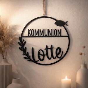 Könnte beinhalten: Schwarzes Metall-Wandschild mit dem Wort "KOMMUNION" über dem Namen "Lotte" in Schreibschrift. Ein Fischsymbol und ein Blättermotiv sind in das kreisförmige Design integriert. Das Schild wird von einer braunen Schnur gehalten.