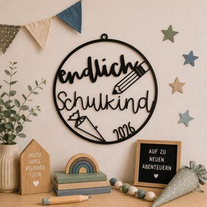 Puede incluir: Un adorno de pared circular negro con el texto en alemán "endlich Schulkind", un icono de lápiz, un icono de cono escolar y el año "2026". El diseño está hecho de un material plano e incluye un pequeño lazo en la parte superior para colgar.