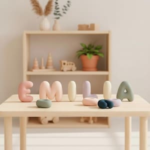 Può includere: Un set di lettere lisce, arrotondate e con finitura opaca che compongono il nome EMILIA in tenui colori pastello tra cui rosa, pesca, crema, lavanda e verde salvia, accompagnate da piccoli oggetti arrotondati a forma di anello.