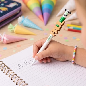 Könnte beinhalten: Ein weißer Kugelschreiber mit Panda-Aufsatz, dem Namen PAUL aus Holzbuchstaben und dem in Grün aufgedruckten Text HUG ME. Der Stift wird verwendet, um die Buchstaben ABC in ein liniertes Spiralnotizbuch zu schreiben, umgeben von bunten Papierkegeln.