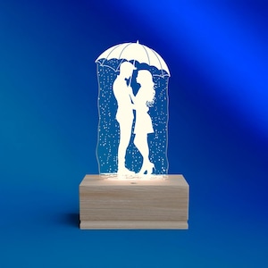 May include: A decorative lamp featuring a silhouette of a couple under an umbrella in the rain. The white figures are set against a blue background, with the lamp resting on a wooden base. The lamp is illuminated.