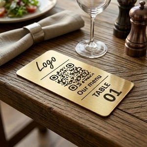 May include: A rectangular gold-colored table sign with a QR code and the words "Logo", "Our menu", and "TABLE 01" printed in black. The sign is on a wooden table, next to a wine glass, napkin, and salt and pepper shakers.