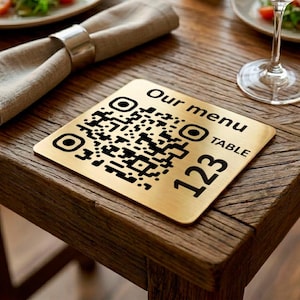 May include: A square gold-colored metal sign with rounded corners featuring a black printed QR code, the text "Our menu", the word "TABLE", and the number "123". The sign has a brushed metallic finish and is designed for display on a table.
