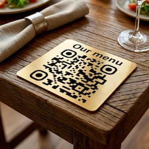 May include: A square, gold-colored QR code coaster with the words "Our menu" printed on it. The coaster is on a wooden table, next to a napkin and a wine glass. The QR code is black.