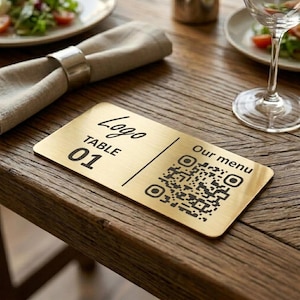 May include: A rectangular gold table sign with the word "Logo" in cursive, "TABLE 01", and a QR code. The words "Our menu" are printed next to the QR code. The sign is on a wooden table, with a napkin and a wine glass in the background.