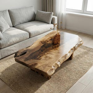 May include: A natural wood coffee table with a live edge design. The table has a light brown finish with darker wood grain patterns. It sits on a rectangular base and is placed on a woven rug in front of a gray sofa.