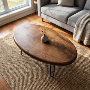 Peut inclure: Table basse ovale en bois avec une finition marron foncé et des pieds épingles noirs. Un petit vase avec des fleurs séchées est posé sur la table. La table est posée sur un tapis en jute tressé dans un salon.