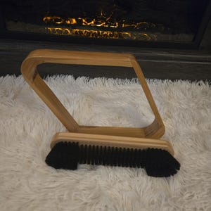 May include: A wooden pool cue rack with a black brush for cleaning the cue. The rack is made of light-colored wood and has a unique, curved design. The brush is attached to the base of the rack.