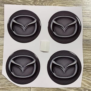 Könnte beinhalten: Vier runde Aufkleber mit dem Mazda-Logo. Jedes Logo ist ein silbernes Emblem auf einem schwarz-grauen strukturierten Hintergrund. Die Aufkleber befinden sich auf einem weißen Blatt Papier.