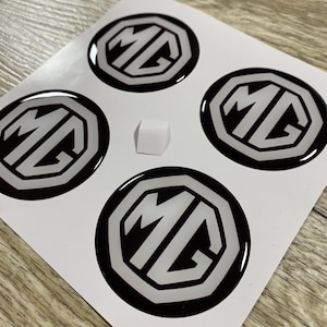 May include: Four black and white MG logo stickers on a white sheet. Each sticker is a circle with a black border and the white letters "MG" inside. A small white square is between the stickers.