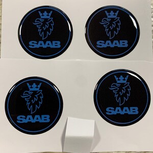 Könnte beinhalten: Vier schwarze und blaue Saab-Emblem-Aufkleber. Jeder Aufkleber ist rund mit einem blauen Saab-Logo und -Text. Die Aufkleber befinden sich auf weißem Hintergrund.