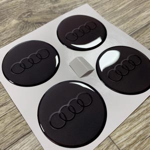 Op de afbeelding: Vier zwarte, ronde auto-embleemstickers met het Audi-logo, gerangschikt op een witte achtergrond. De stickers hebben een glanzende afwerking en een verhoogd ontwerp. Er is ook een klein, grijs, rechthoekig stuk aanwezig.