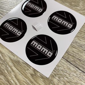 May include: Sheet of four black and white MOMO logo stickers. Each sticker is a black circle with the word "MOMO" in white, and a white arrow design. A small white square is also visible.