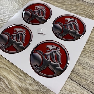 May include: Four circular stickers with a silver lion emblem set against a red backdrop. The stickers are on a white sheet, accompanied by a small, white square sticker. The stickers have a glossy finish.