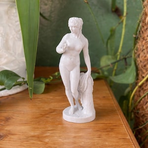 Może przedstawiać: Biała marmurowa statua nagiej postaci kobiecej, stojąca na okrągłej podstawie. Statua trzyma w rękach kawałek materiału i przedmiot. Statua ma około 15 cm wysokości.