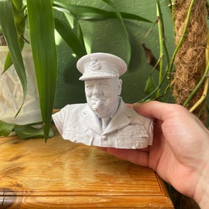 Peut inclure: Buste blanc de Winston Churchill, le représentant en uniforme militaire avec un chapeau et un cigare. La sculpture est tenue dans une main, avec une surface en bois et des plantes vertes en arrière-plan.