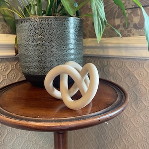 May include: A matte off-white decorative object featuring a continuous, interlocking knot design with smooth, rounded surfaces. The sculpture is composed of a single, fluid loop that twists into a complex, three-dimensional geometric shape.