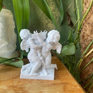 Puede incluir: Estatua blanca de querubines con dos figuras sosteniendo una cruz. Los querubines están arrodillados sobre una base rectangular. La estatua está hecha de un material moteado, con plantas verdes de fondo.